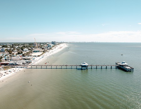 An aerial view of a coastal area with a long pier extending into the ocean, a sandy beach, and buildings along the shoreline ending the sentence.