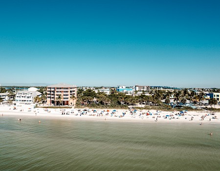 The image shows a coastal scene with a sandy beach, people relaxing under umbrellas, residential buildings in the background, and calm green waters.