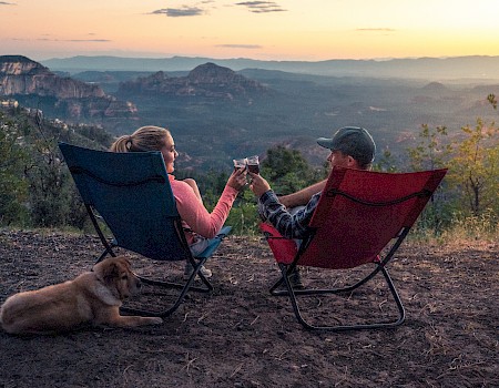 Two people sit on camping chairs, toasting with drinks while a dog sits beside them, overlooking a scenic mountainous landscape at sunset.