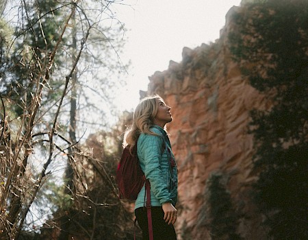 A person with a backpack stands outside, looking up at a rocky, cliff-like structure, surrounded by trees and nature. The scene is serene and contemplative.