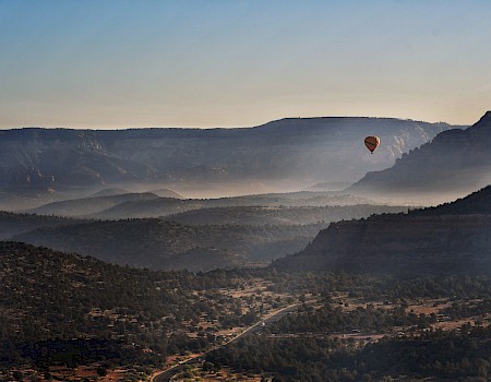 A hot air balloon floats over a scenic, mountainous landscape with mist settling in the valleys and the morning sun casting soft light.