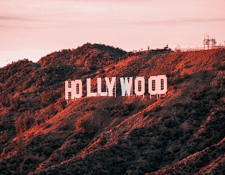 The image showcases the iconic Hollywood sign situated on a hillside, with a backdrop of rugged terrain and a soft pink-hued sky, ending the sentence.