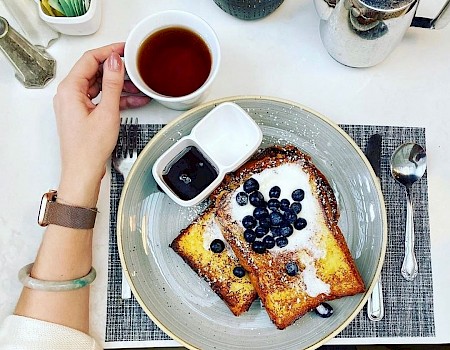 A person is having breakfast with toast topped with blueberries, powdered sugar, and syrup, a cup of tea, and various condiments on a table.