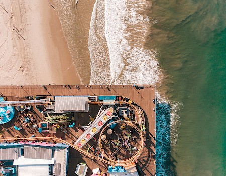 An aerial view of a seaside amusement park with roller coasters and rides on a pier, beside a beach and the ocean.