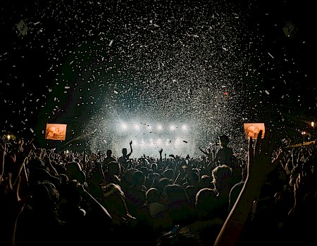 The image shows a concert crowd with raised arms and confetti falling from above. Bright stage lights and screens are visible in the background.