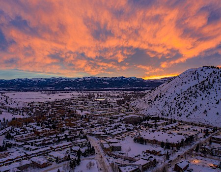 A snowy town is nestled between hills, illuminated by a dramatic, colorful sunset sky filled with vibrant orange and pink clouds.