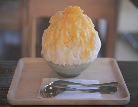 The image shows a bowl of shaved ice topped with mango pieces, placed on a tray with two spoons and a napkin.