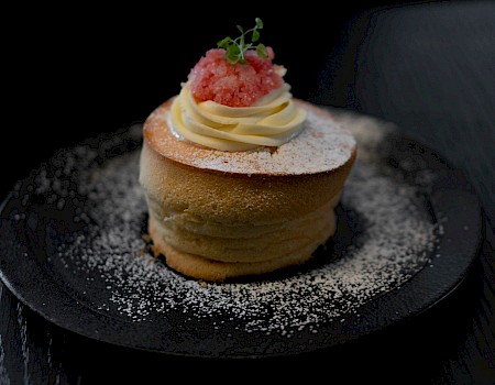 A beautifully plated dessert, likely a fluffy pancake or cake, topped with cream, pink granita, and a sprig of herbs, dusted with powdered sugar.