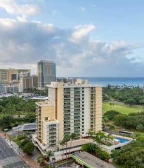 The image shows a cityscape with tall buildings, a road, green parks, and a view of the ocean under a partly cloudy sky.