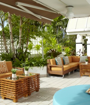 A stylish outdoor patio features orange-brown sofas with patterned cushions, wooden tables, and lush green plants set against a tiled floor.